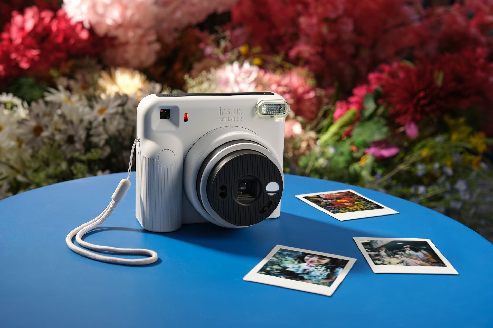 Bílý Fujifilm instax SQUARE 2 s vytištěnými čtvercovými fotografiemi mezi květinami
