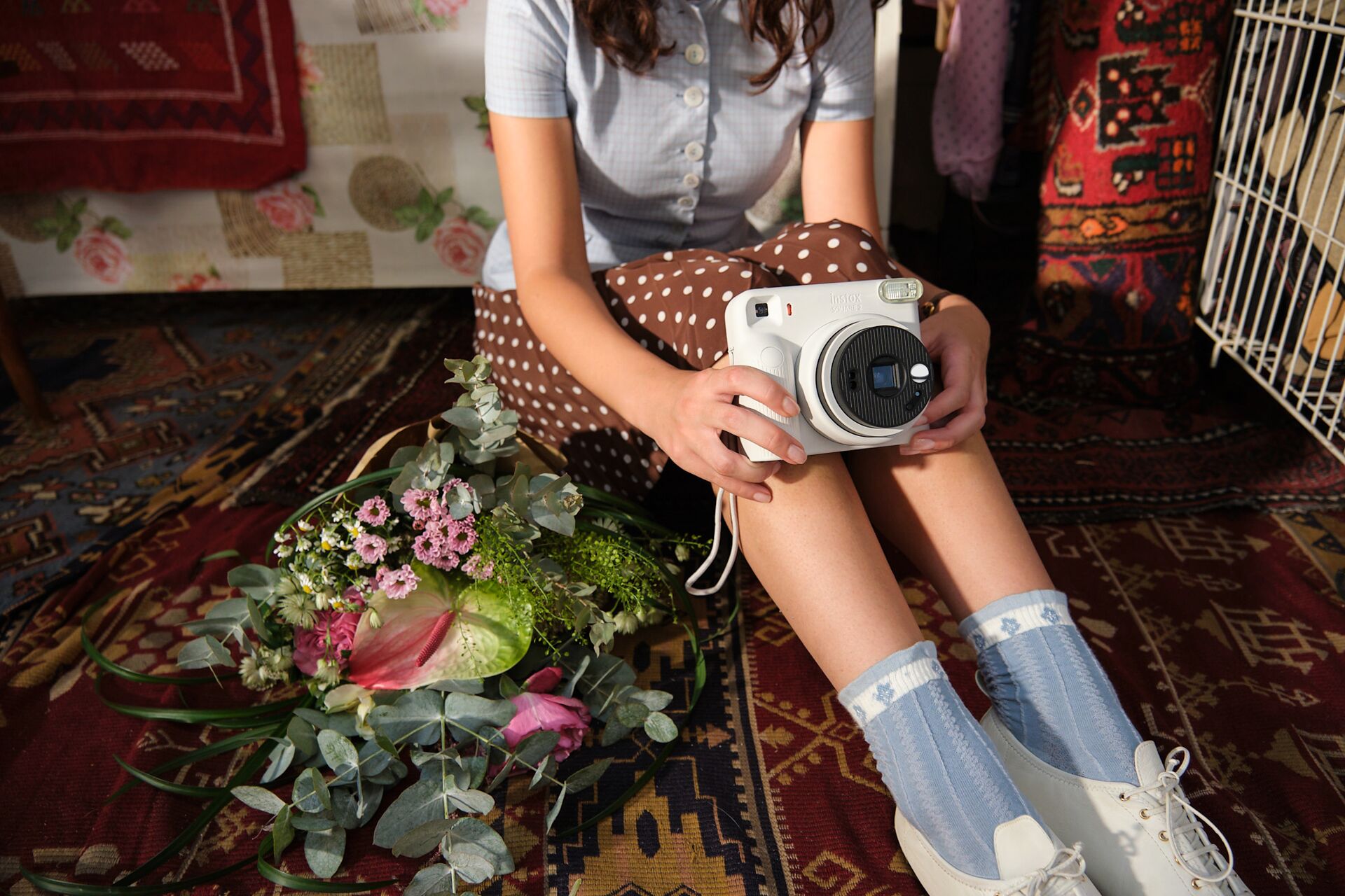 Žena drží bílý instantní fotoaparát Fujifilm instax SQUARE 2 v domácím prostředí